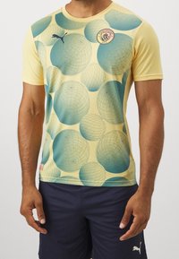 Krátký rukáv, žlutá košile s geometrickým vzorem modrých a tyrkysových kruhů, s logem Puma a znakem Manchester City na hrudi.