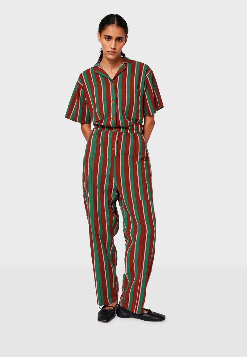 Žena v krátkom rukáve tričku s hnedými, zelenými a béžovými vertikálnymi pruhmi a nohavicami, stojí s rukami za chrbtom, čierne baleríny.