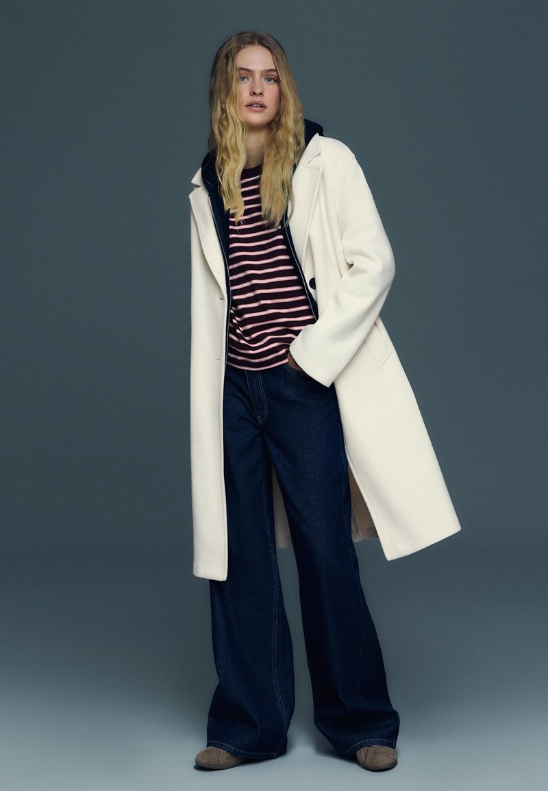 Femme aux longs cheveux blonds portant un manteau blanc par-dessus un chemisier rayé rose et noir, un jean large foncé et des chaussures marron.