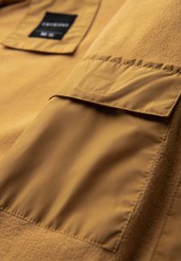 Gros plan d'une veste marron montrant une poche en tissu avec un rabat et une étiquette rectangulaire noire sur la partie supérieure.