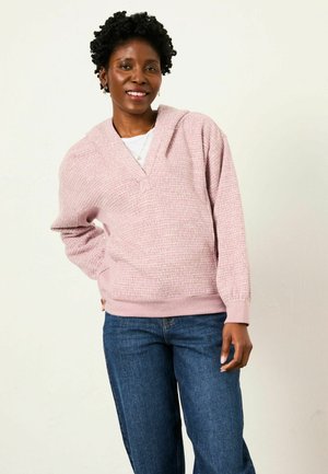 Pink textured hoodie with a V-neck and wide sleeves, paired with blue jeans. The fabric appears soft and warm, featuring a relaxed fit.