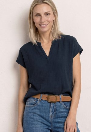Uśmiechnięta blondynka, ubrana w granatową bluzkę z krótkim rękawem włożoną w niebieskie dżinsy z brązowym skórzanym paskiem, stoi na tle jasnej ściany.