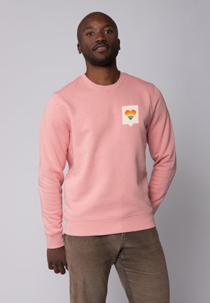 Lichtroze sweatshirt gemaakt van zacht materiaal met een hartlogo met regenboogstrepen op een witte spraakballonpatch. Ronde halsontwerp.