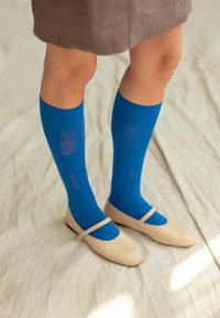Calcetines azules hasta la rodilla combinados con zapatos planos beige con tiras, usados con una falda marrón. La superficie es lisa y uniforme.