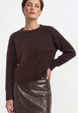 Woman wearing a dark brown fuzzy sweater and a shiny brown snakeskin-patterned skirt with her hand on her hip against a white background.