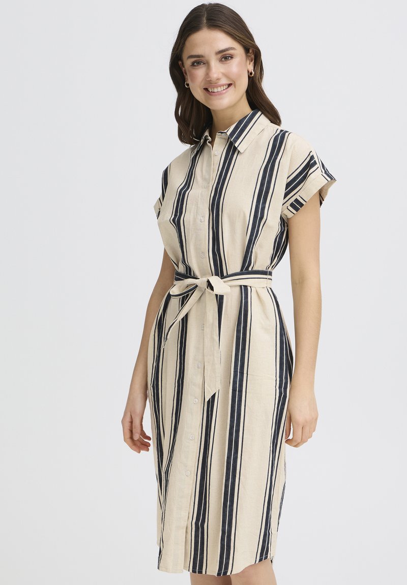 Femme souriante, portant une robe boutonnée beige à rayures verticales noires avec une ceinture nouée à la taille, debout devant un fond uni.