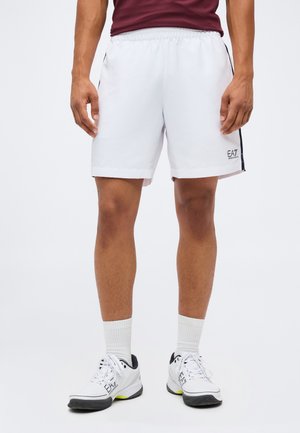 Mann trägt weiße Sportshorts mit schwarzen Seitenstreifen und passende weiße Sportschuhe mit Socken vor einfarbigem Hintergrund.