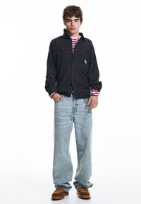 Zwart geruit overhemd met knoopsluiting, gedragen over een gestreept roze shirt. Lichtblauwe spijkerbroek met geborduurde details en bruine schoenen.