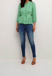 Donna che indossa un top a maniche lunghe verde a fantasia, jeans skinny blu e sandali con tacco marroni, in piedi su uno sfondo bianco semplice.