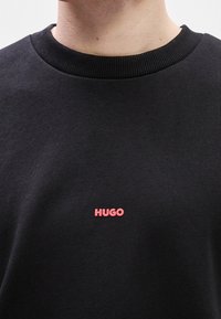 Gros plan d'une personne portant un sweat-shirt noir à col rond avec un petit texte rouge "HUGO" brodé au centre de la poitrine.
