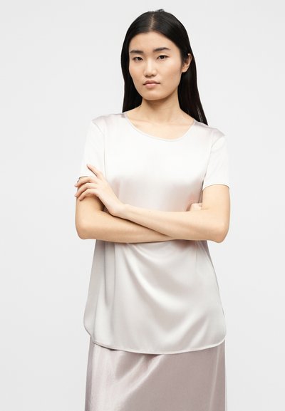 Νεαρή γυναίκα με μακριά σκούρα μαλλιά, φορώντας ένα ανοιχτό ροζ σατέν μπλουζάκι με κοντά μανίκια και αντιστοιχισμένη φούστα, στέκεται με σταυρωμένα χέρια.