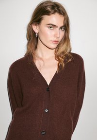 Brun stickad cardigan med V-hals, prydd med tre svarta knappar fram. Mjukt material och avslappnad passform.