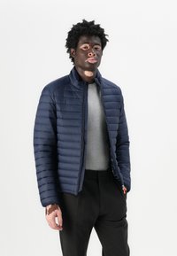 Veste en duvet bleu marine avec un matelassage horizontal, un col haut et une fermeture éclair à l'avant. Portée sur un T-shirt gris avec un pantalon noir.
