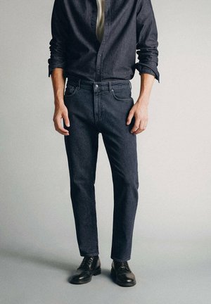 Homme portant une chemise noire boutonnée, un jean en denim foncé et des chaussures habillées noires, debout devant un fond clair uni.
