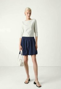 Λευκή, μακριά μπλούζα συνδυασμένη με μια μπλε, ραμμένη φούστα. Τα αξεσουάρ περιλαμβάνουν μια μικρή λευκή τσάντα και σανδάλια με λουριά, με φόντο ουδέτερη επιφάνεια.