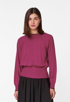 Magenta Langarm-Pullover mit ribbeltem Taillenbund und Bündchen, im entspannten Schnitt und mit Rundhalsausschnitt. Darunter ein schwarzer Rock.