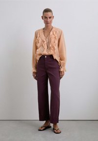 Mujer vestida con una blusa de volantes color durazno, pantalones morados de cintura alta, sandalias negras y un collar largo, de pie contra una pared clara.