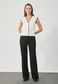 Fehér ujjatlan felső fekete díszítéssel, V-nyakkivágással és gombos záródással, fekete bővített szárú nadrággal és magassarkú cipővel párosítva.