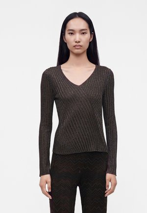 Femme aux longs cheveux noirs portant un haut marron foncé ajusté à manches longues à côtes diagonales et un pantalon noir à motifs sur fond blanc.