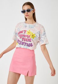Tie-dye t-shirt med korte ærmer med multifarvet blomstergrafik og tekst; parret med en pink mini-skjørt med en sideslids. Blå rektangulære solbriller.