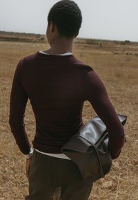 Tummanruskea nahkalaukku, jossa on laskostettu läppä ja hihna, pidetään marooninvärisen pitkähihasen paidan vieressä, kuivassa, ruohikkoisessa maisemassa.