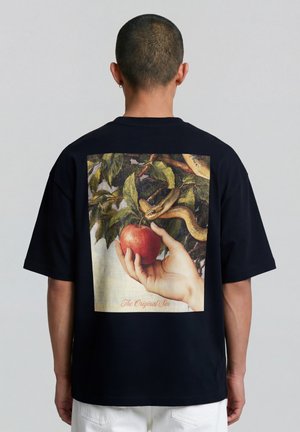 Man met een zwart shirt met een afbeelding van een hand die een rode appel vasthoudt, een slang die zich om takken kronkelt erboven, en de tekst "The Original Sin" op de achterkant.