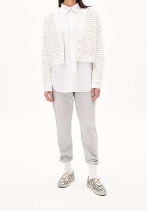 Femme portant une chemise blanche boutonnée, un cardigan court en maille crème, un pantalon de jogging gris, des chaussettes blanches et des baskets à imprimé zèbre, debout sur fond blanc.