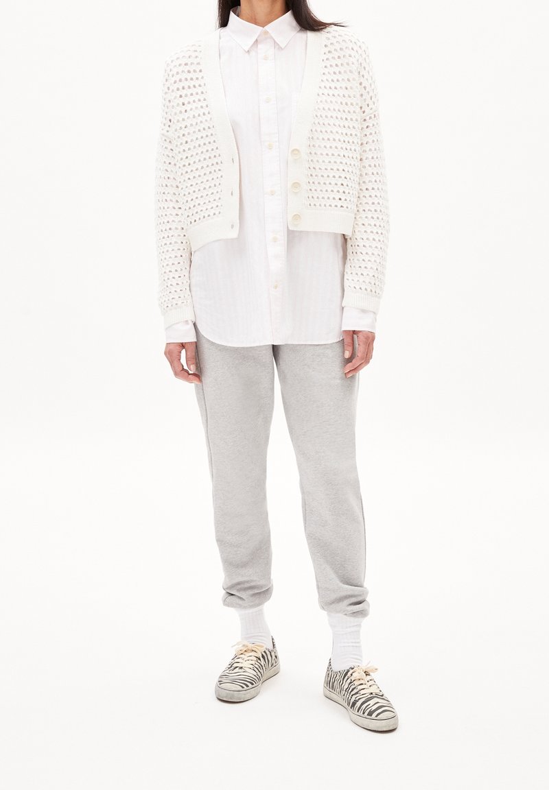 Femme portant une chemise blanche boutonnée, un cardigan court en maille crème, un pantalon de jogging gris, des chaussettes blanches et des baskets à imprimé zèbre, debout sur fond blanc.