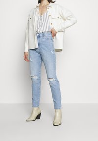 Veste blanche boutonnée, haut rayé gris et blanc, jean bleu clair déchiré, et bottines beiges à motif avec un talon épais.