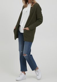 Olivově zelený pletený kardigan s otevřeným předním dílem, dlouhými rukávy a kapsami, nošený přes bílou košili, v kombinaci s roztrhanými modrými džíny a bílými teniskami.