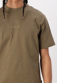 Olive zelené bavlnené tričko s krátkymi rukávmi, s vyšitým logom "REPLAY" v béžovej farbe na hrudi. Hladká textúra.