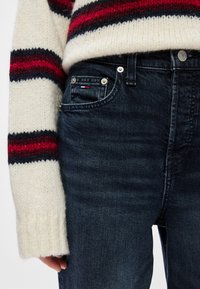 Pulover tricotat cu dungi orizontale roșii și bleumarin, asortat cu blugi de denim de culoare albastră închis, care au o uniune cu un mic logo roșu, alb și albastru.