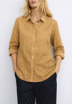 Donna che indossa una camicia mostarda a maniche lunghe con bottoni e maniche arrotolate, abbinata a pantaloni scuri a righe sottili, con le mani in tasca.