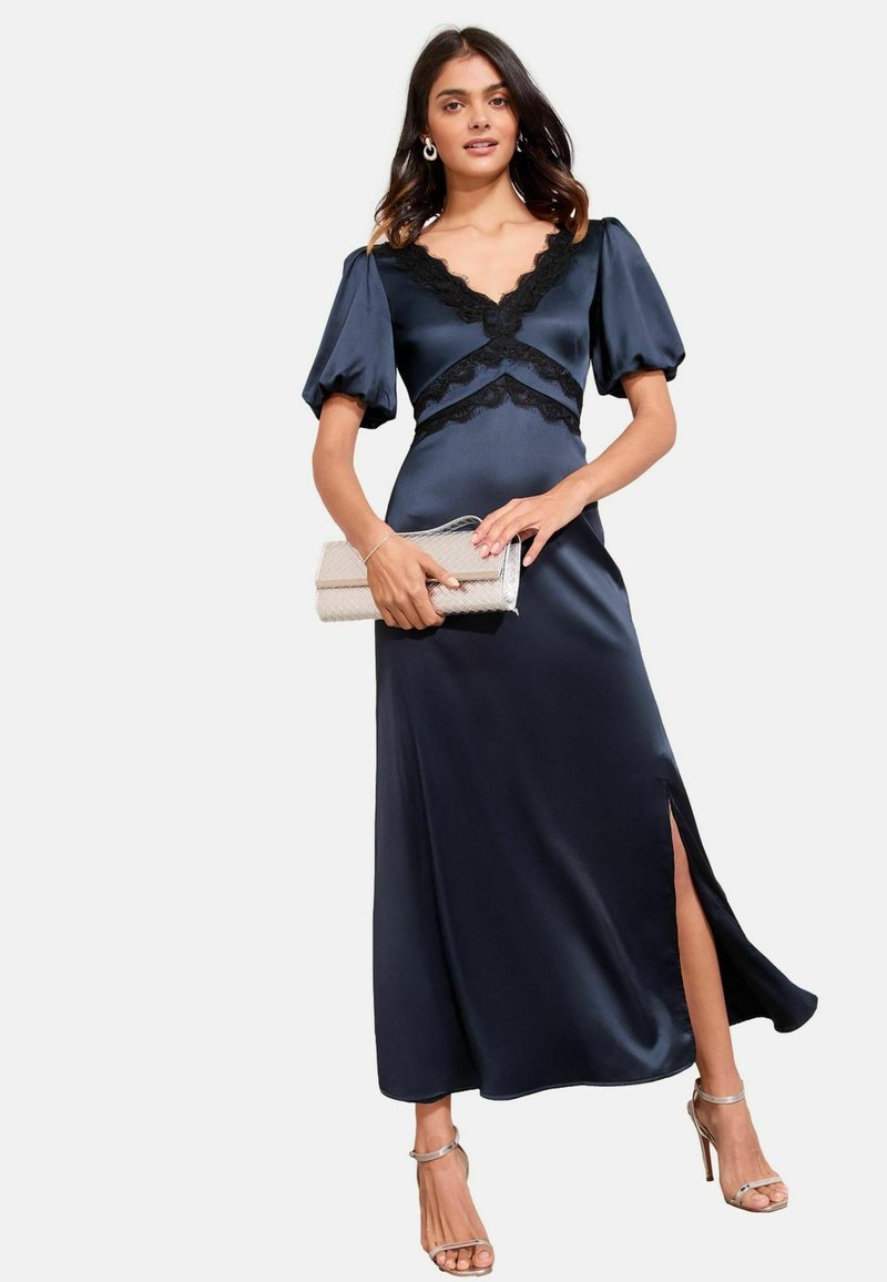 Navyblaues Satinkleid in langer Ausführung mit Spitzenbesatz, Puffärmeln und seitlichem Schlitz. Kombiniert mit einer metallicfarbenen Clutch und Riemchen-High Heels.