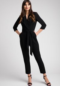 Czarny kombinezon z fasonem kopertowym, z rękawami o długości 3/4, pasek w talii i zwężanymi nogawkami, w połączeniu z czarnymi sandałami na obcasie.