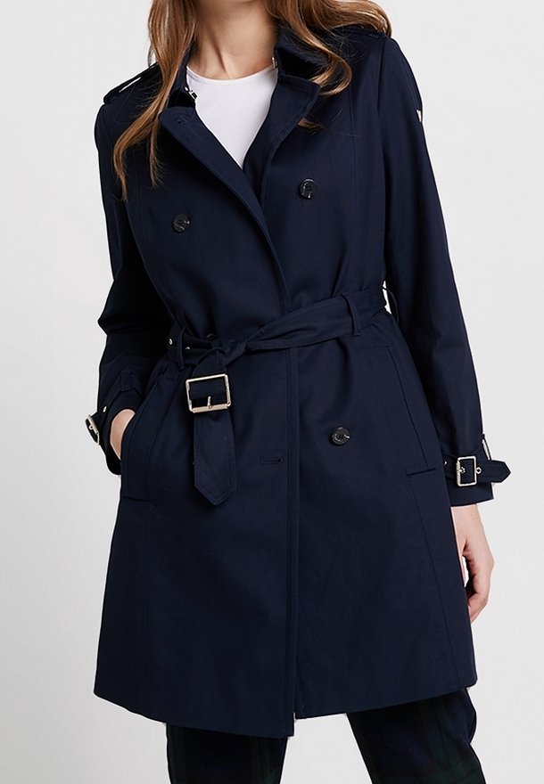 Femme portant un trench-coat bleu marine à double boutonnage avec ceinture à la taille et poignets à boucles par-dessus un haut blanc.