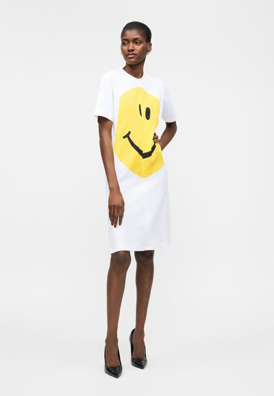 Femme portant une robe blanche mi-longue avec un grand visage souriant jaune, debout avec la main sur la hanche, talons noirs à bout pointu, fond neutre.