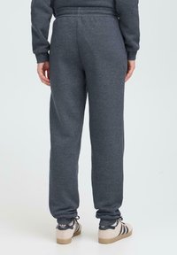Person som har på seg mørkegrå joggebukser og beige sko med svarte striper, står på en lys bakgrunn, sett fra bak.