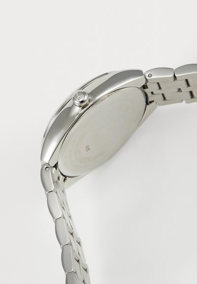 Zilveren roestvrijstalen horloge met een ronde kast, getextureerde kroon en een schakelsluiting. Het gladde bandje gaat over in een platte achterzijde van de kast.
