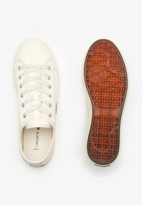 Zapatillas de lona blancas con ojales plateados, cordones planos y una suela de goma marrón con un patrón que presenta un logo en el lateral. Parte superior texturizada.