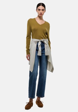 Pull en maille vert olive à col en V, chemise légère rayée nouée à la taille, jean bleu à jambes larges, et chaussures en suède marron à lacets.