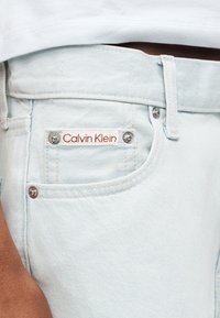 Τσέπη από ανοιχτό μπλε τζιν με ετικέτα "Calvin Klein" ραμμένη πάνω από τα καρφιά, φορεμένη με λευκό τοπ.