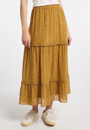 Kvinde iført et sennepsgult blomstret flæset midi-nederdel, hvide sneakers og en hvid, indstoppet top, stående på en hvid baggrund.