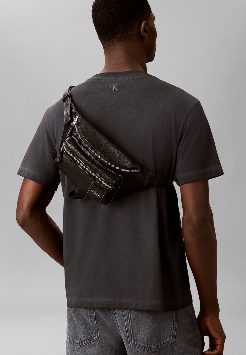 Homme portant un t-shirt gris foncé et un jean avec un sac bandoulière en cuir noir porté dans le dos.
