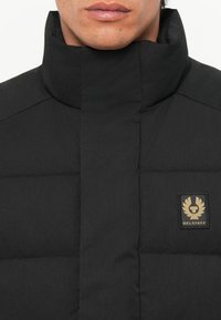 Black padded jacket with high collar, featuring a gold emblem patch. The fabric has a smooth texture with subtle seams and stitching details.