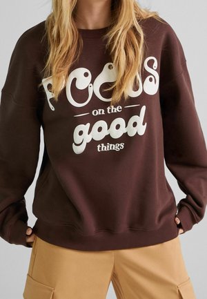 Personne portant un sweat-shirt marron avec le texte « Focus on the good things », assorti à un pantalon beige. Mains partiellement dans les poches.