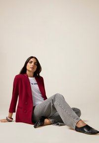 Blazer burdeos de doble botonadura sobre una camiseta gráfica blanca, jeans grises y bailarinas negras decoradas con tachuelas. Tejido suave y corte entallado.