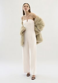 Tuta senza spalline di colore rosa chiaro con dettaglio a chiave frontale, indossata con un cappotto in pelliccia sintetica beige. Tacchi aperti neri completano il look.