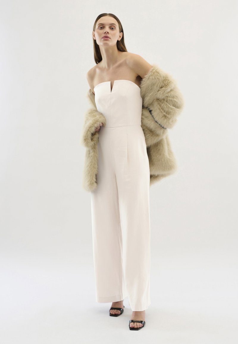 Tuta senza spalline di colore rosa chiaro con dettaglio a chiave frontale, indossata con un cappotto in pelliccia sintetica beige. Tacchi aperti neri completano il look.