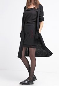 Schwarzer langer Cardigan über einem schwarzen Oberteil und Rock mit floralem Design. Schwarze Strumpfhosen und Schuhe vervollständigen das Outfit.